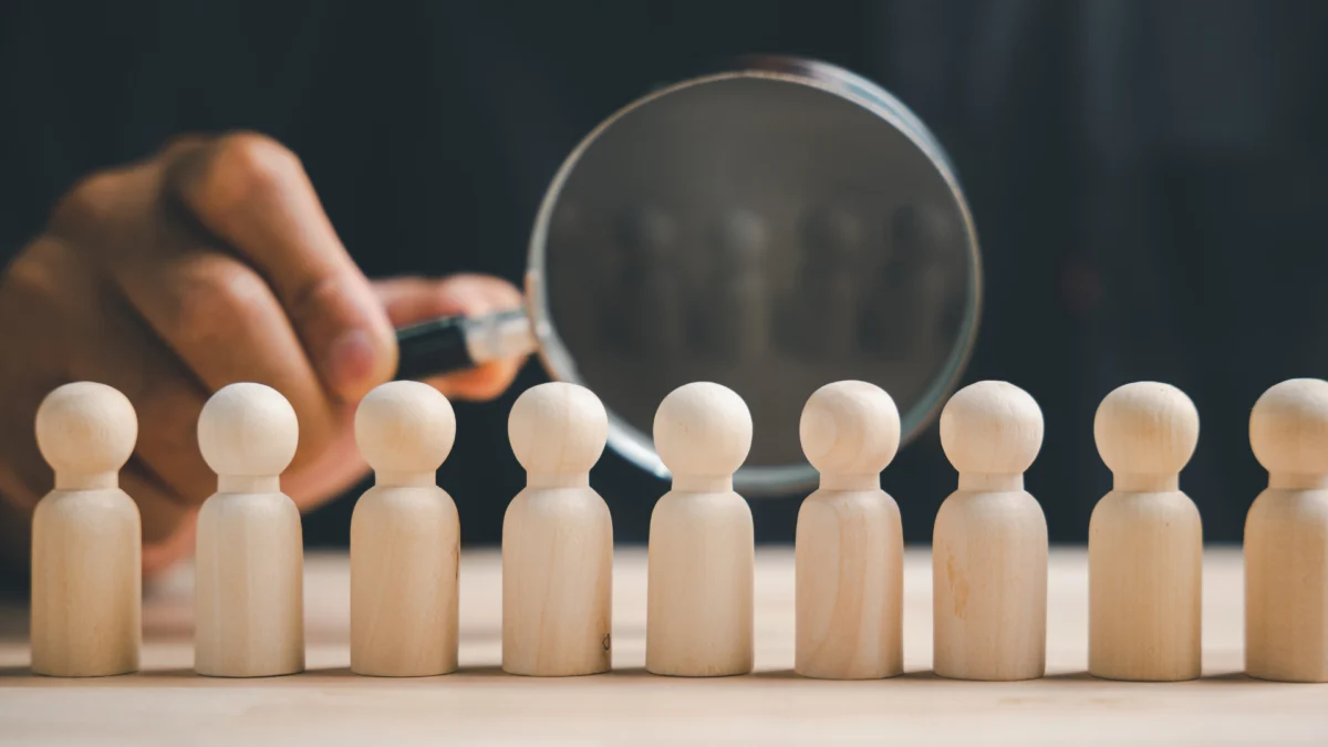 Mão segurando uma lupa sobre bonecos de madeira alinhados, simbolizando a análise de mercado e a definição de público-alvo em um plano de negócios para água mineral. A imagem representa a seleção estratégica de nichos de envase premium ou de volume.